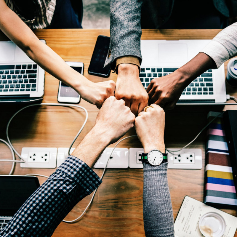 Five people fist-bumping over their work station and laptops