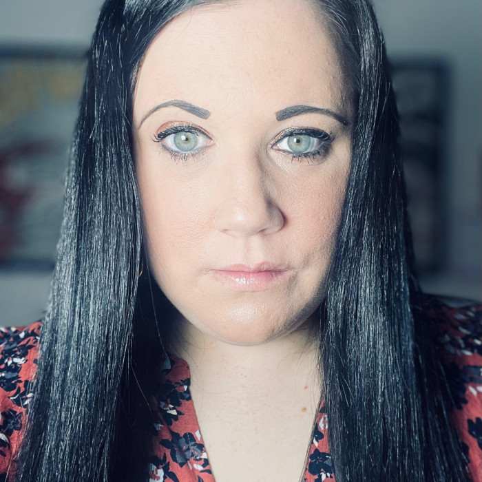 Louisa looking forward in front of a plain background. Louisa has brown hair and is wearing a blue top